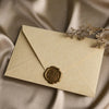 A pale yellow linen texture envelope with a gold sealing wax sticker rests on draped beige fabric.