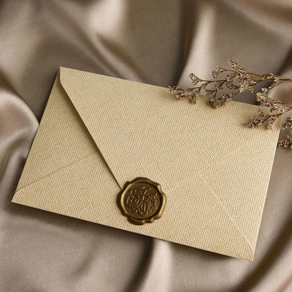 A pale yellow linen texture envelope with a gold sealing wax sticker rests on draped beige fabric.