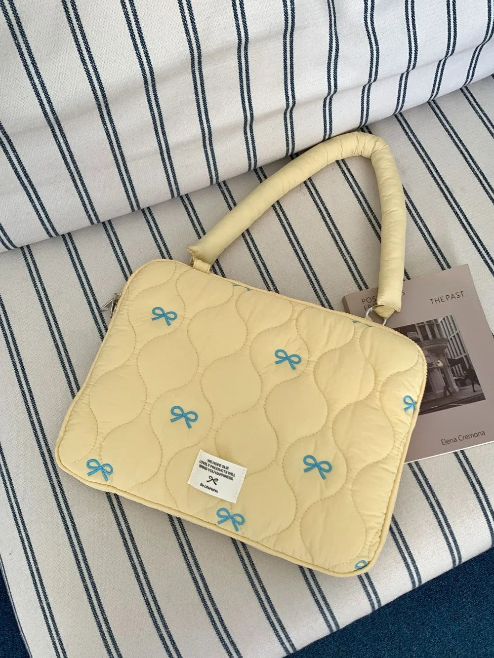 A light yellow quilted laptop shoulder bag with a leather handle and blue bow embellishments resting on striped fabric.