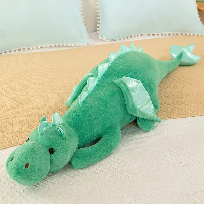 A soft, bright green dragon plush toy lying on a beige surface with light blue pillows in the background.