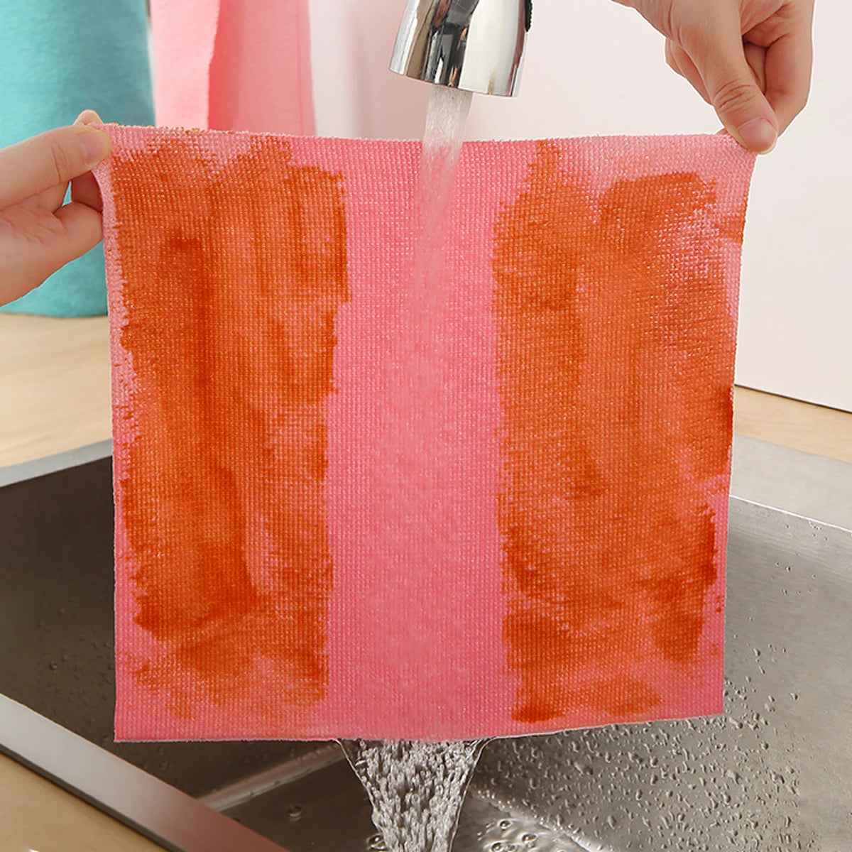 A hand is rinsing a brightly colored orange microfiber cleaning cloth under running water.