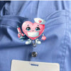 A pink heart-shaped nurse character charm with a stethoscope and ID clip attached to a blue uniform shirt pocket.