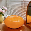 An orange, spherical modern LED wall sconce with a smooth finish sits on a wooden surface next to pink roses.