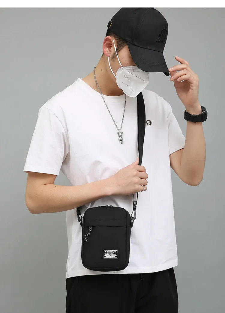 A person wearing a white t-shirt and black cap holds a black mini crossbody phone bag.