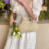 A woman holds a woven straw chain crossbody handbag while standing near floral arrangements.