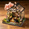 A detailed miniature Japanese sakura tea house model featuring pink blossoms, greenery, and a wooden structure.