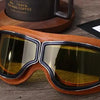 A pair of brown leather retro foldable motorcycle goggles resting on a wooden surface next to a coffee cup.