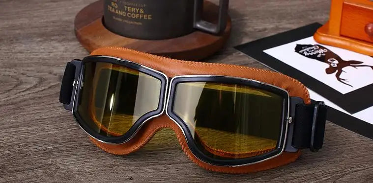 A pair of brown leather retro foldable motorcycle goggles resting on a wooden surface next to a coffee cup.