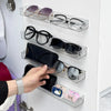 A hand places sunglasses and small accessories into a clear acrylic wall-mounted display shelf.