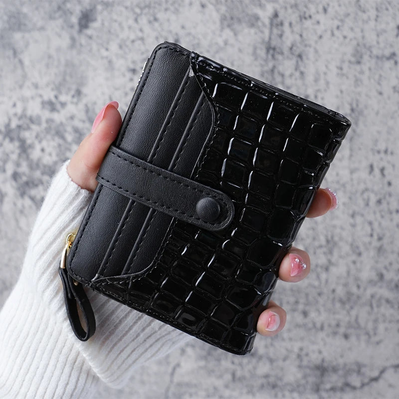 A hand holds a black crocodile-embossed clutch wallet with a stitched design against a gray background.