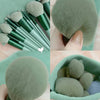 A collection of 13 makeup brushes in various shades of green and gray displayed next to a travel bag.