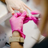 A hand wearing a pink disposable glove is holding a purple fabric, with another hand adjusting a ring on the wrist.
