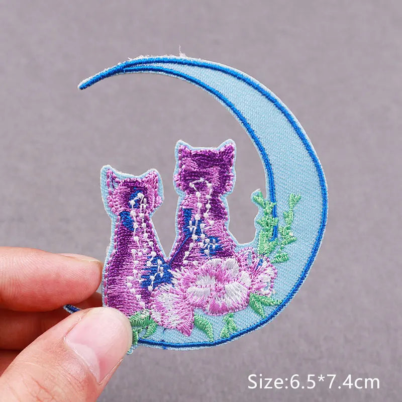 A light blue crescent moon embroidered with two purple floral cat figures on a fabric patch.