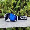 A pair of black and white HD Motocross goggles with a blue anti-fog lens resting on a gray surface amidst green foliage.