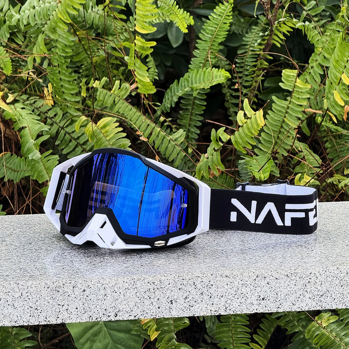 A pair of black and white HD Motocross goggles with a blue anti-fog lens resting on a gray surface amidst green foliage.