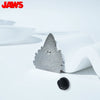 Silver enamel pin shaped like a shark fin, made of zinc alloy, resting on a white surface.