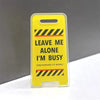 A yellow acrylic desk sign with black hazard stripes that reads Leave me alone I'm busy pretending to work.