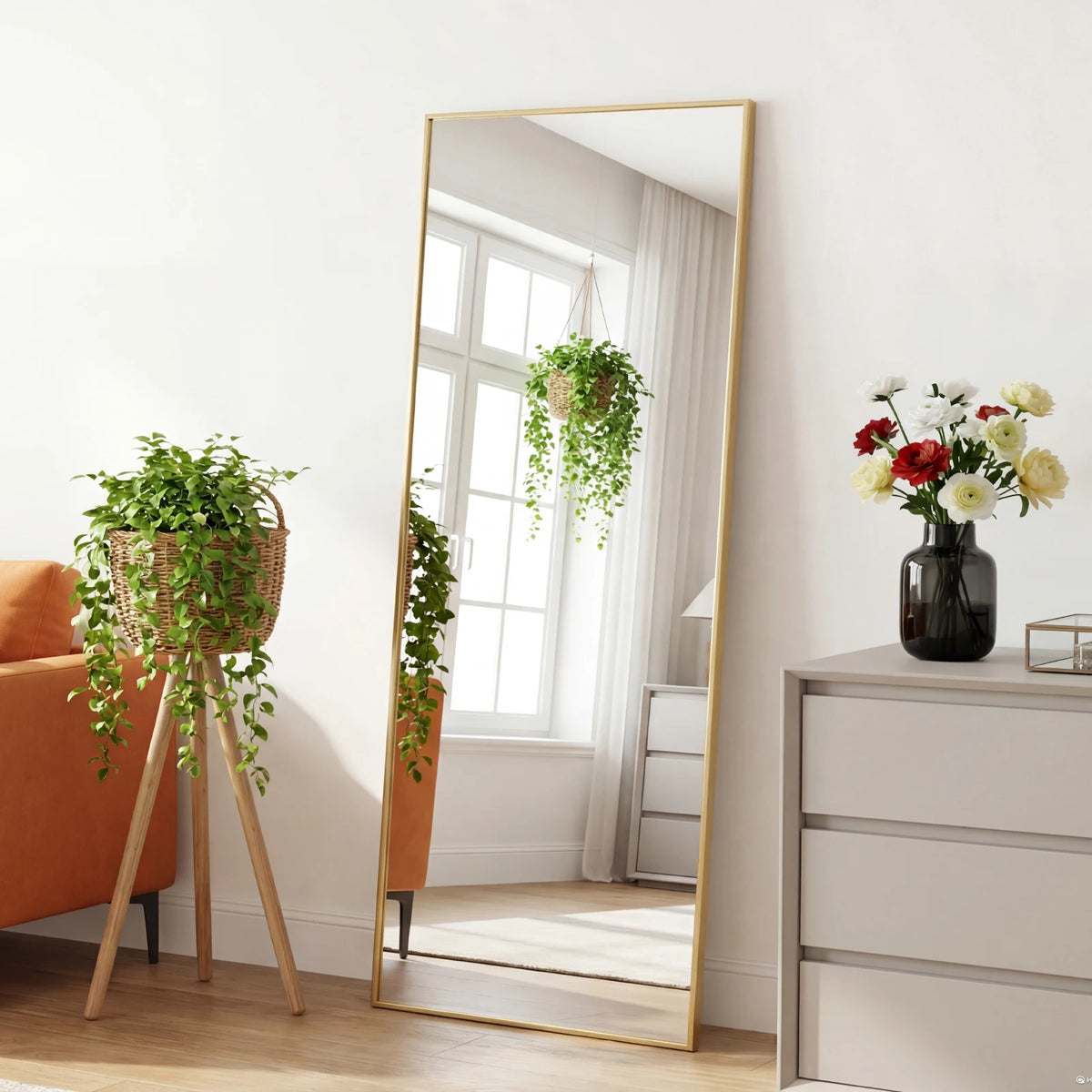 A full length mirror with a gold frame reflects a bright room and greenery, positioned next to an orange sofa.
