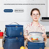 A woman holds a blue insulated lunch bag and an orange fruit on a wooden table.