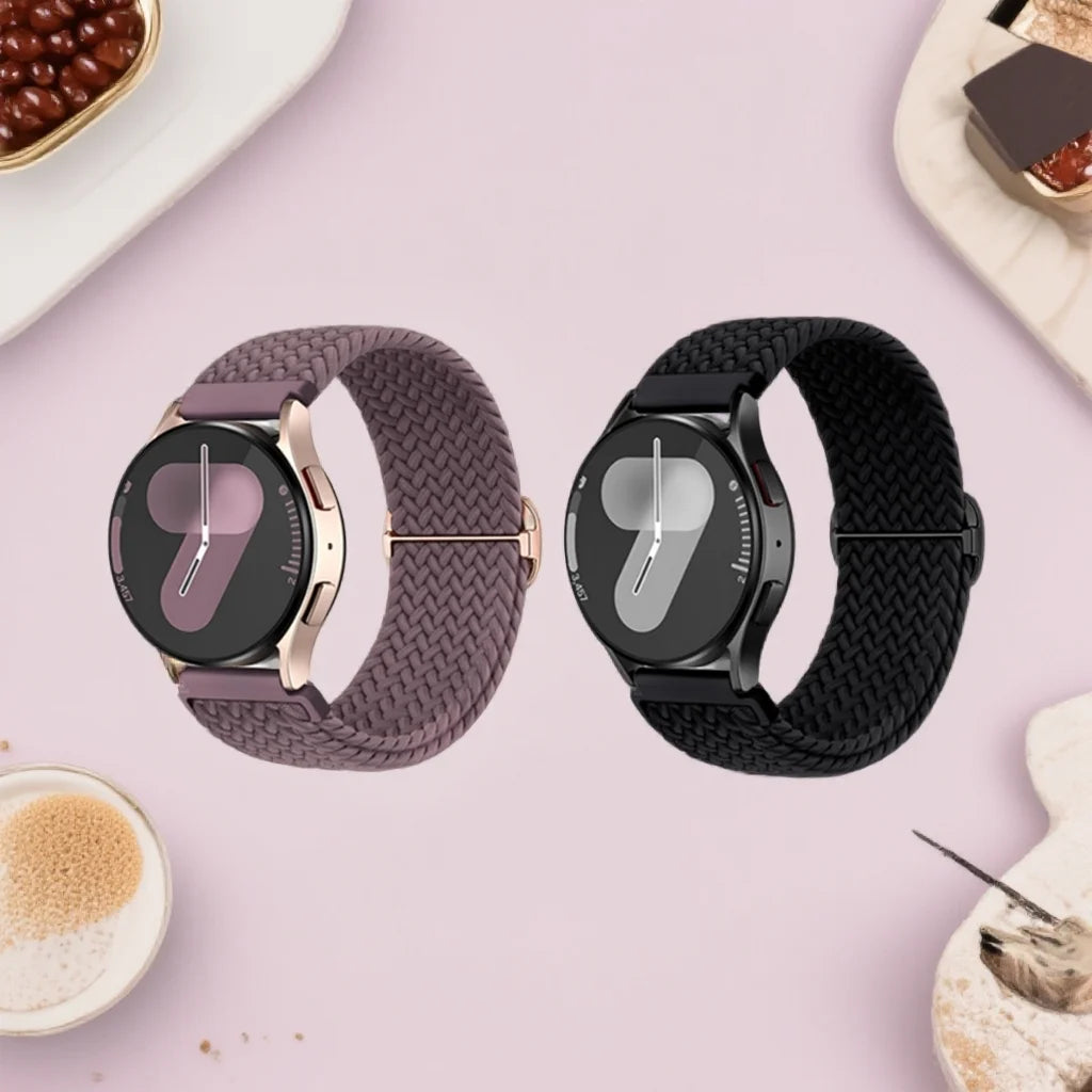A close-up of a purple braided nylon watch band and a black watch with a silver clasp on a pink background.