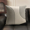 A beige linen cushion cover with black geometric lines is displayed on a dark leather sofa.