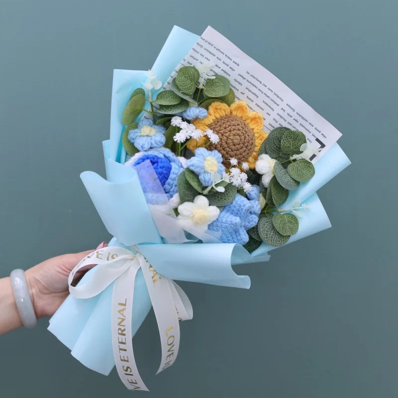Handgefertierter Blumenstrauß aus Häkelwerk mit blauen und gelben Blumen und grünem Laub, in hellblauer Papierverpackung.