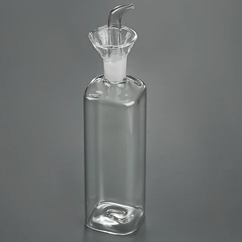 A clear borosilicate glass oil and vinegar dispenser with a stopper and spout, shown against a gray background.