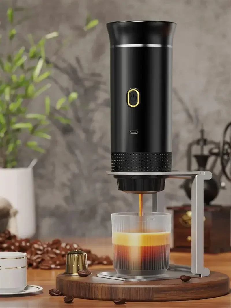 A black portable electric espresso coffee maker is shown on a wooden surface with coffee beans and a glass cup.