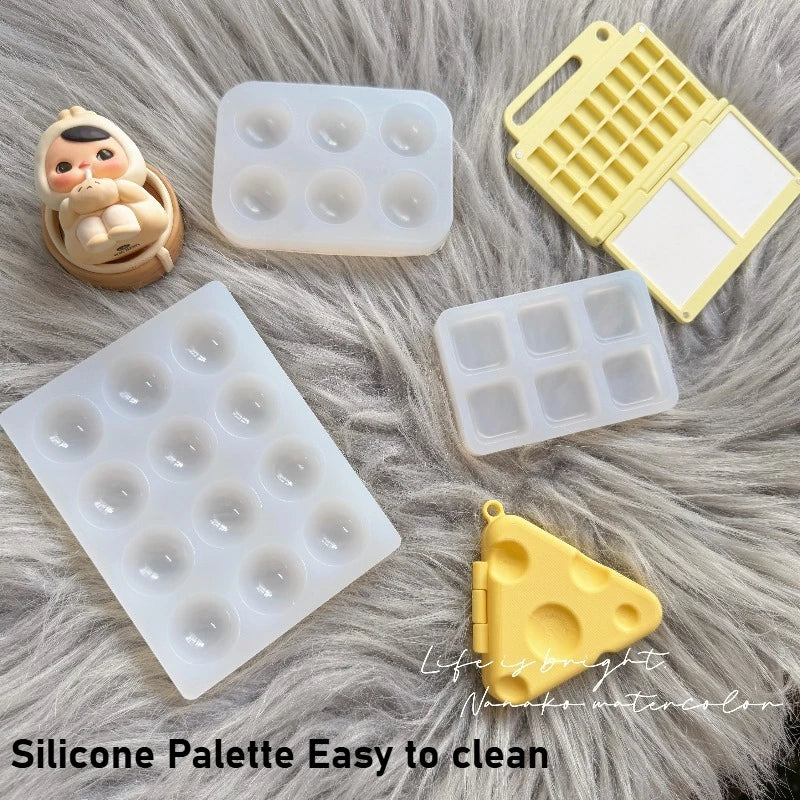 A collection of yellow and white silicone paint palette trays and tools resting on a fluffy gray surface.