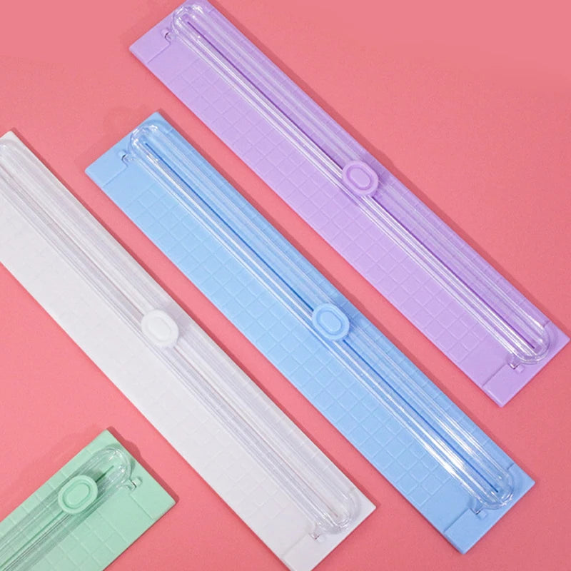 Three portable paper cutters or trimmers in white, light blue, and purple lying on a pink surface.