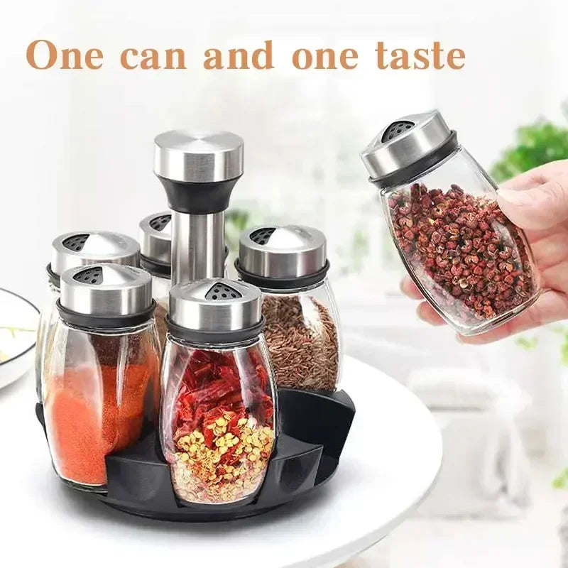 A hand holds a glass spice jar filled with reddish-brown spices, next to a set of rotating glass spice jars.