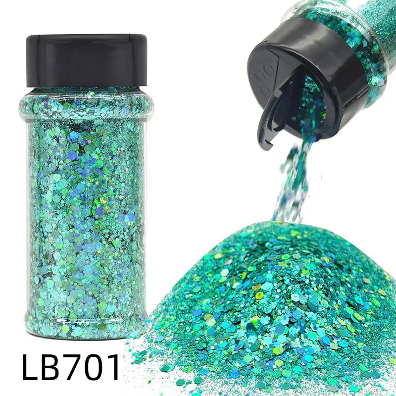 A glass bottle of holographic teal glitter nail mix with a black shaker top and a pile of loose glitter.