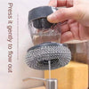 A hand is pressing a dish brush with a built-in soap dispenser to allow liquid detergent to flow out.