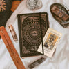 An antiqued tarot deck with meanings on cards is displayed on a white surface next to a wooden stick and other spiritual item