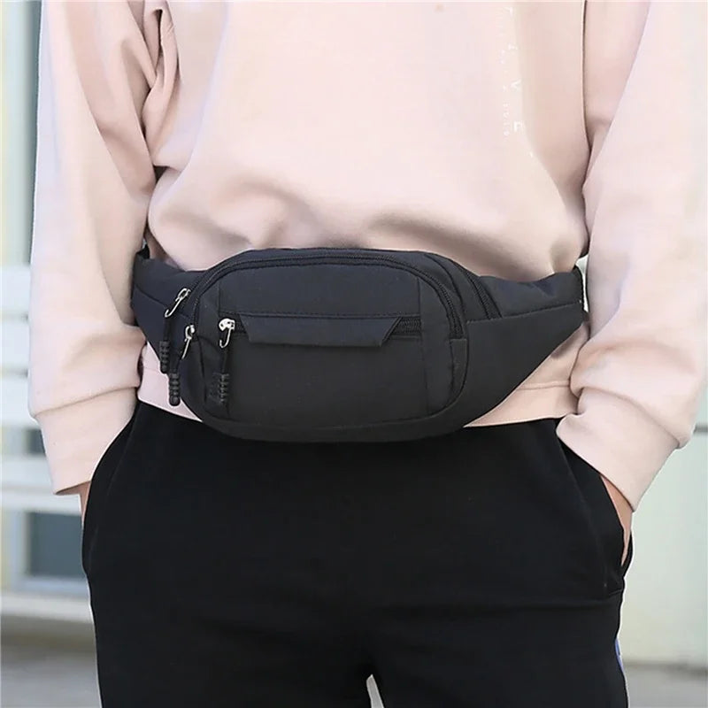A black canvas waist pack with zippers is shown being held by a person wearing a light pink top and black pants.