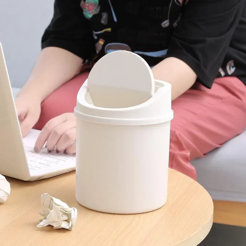 A white compact covered desktop trash bin with a flip lid sitting on a wooden table.
