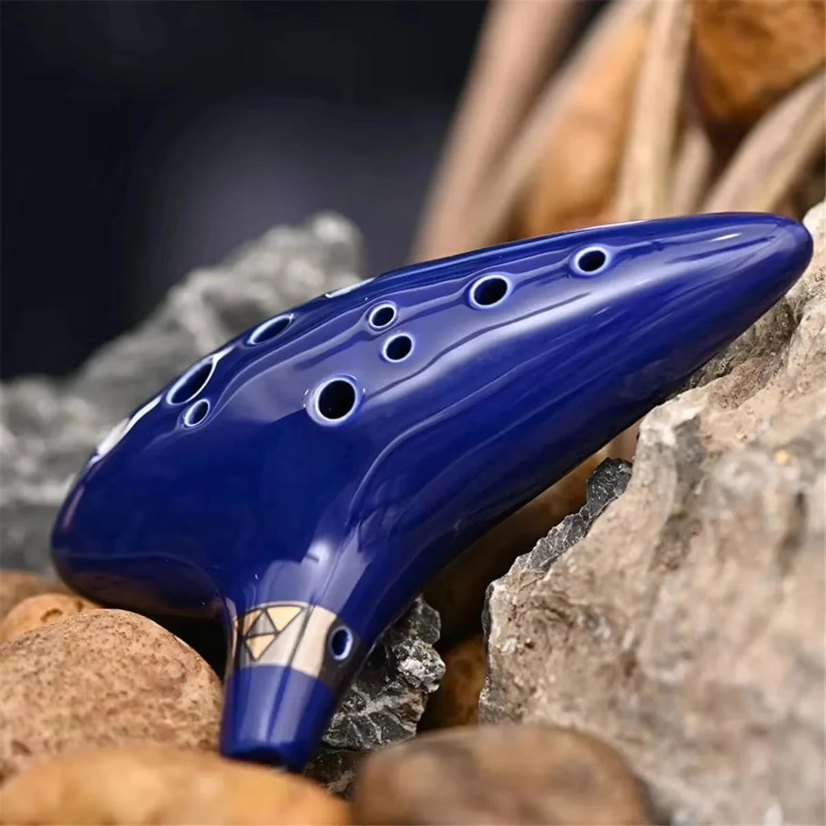 A blue ceramic alto ocarina with holes and a storage bag resting on rocks and dried natural materials.