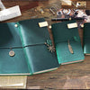 A dark green handcrafted leather refillable journal with brass accents rests on a wooden surface next to paper and tools.