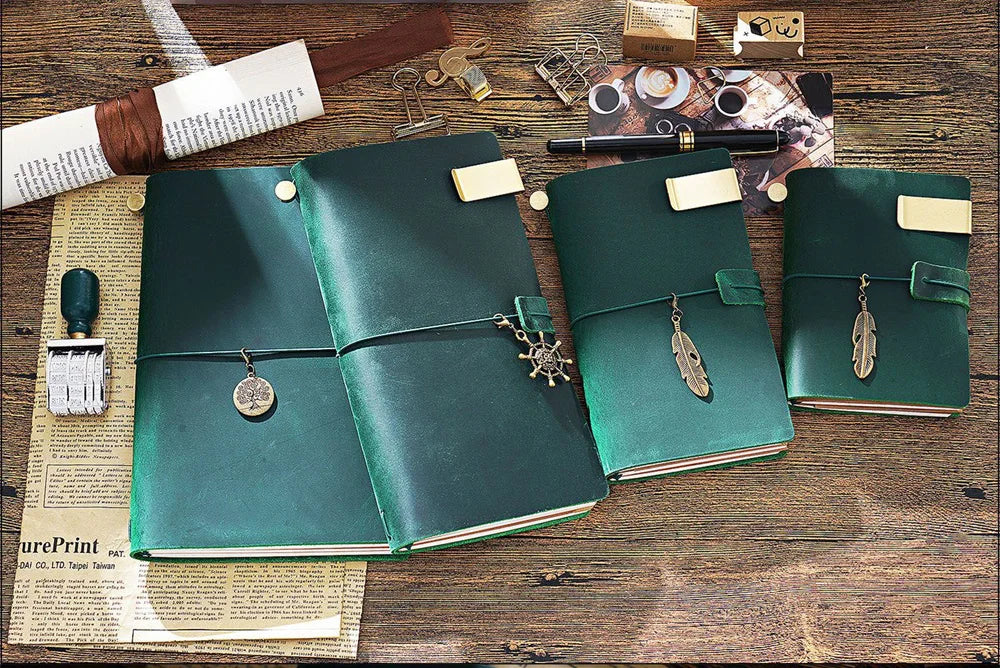 A dark green handcrafted leather refillable journal with brass accents rests on a wooden surface next to paper and tools.