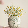 Eine Hand hält eine weiße Keramikvase mit gelben und weißen Wildblumen vor einem hellen Hintergrund.