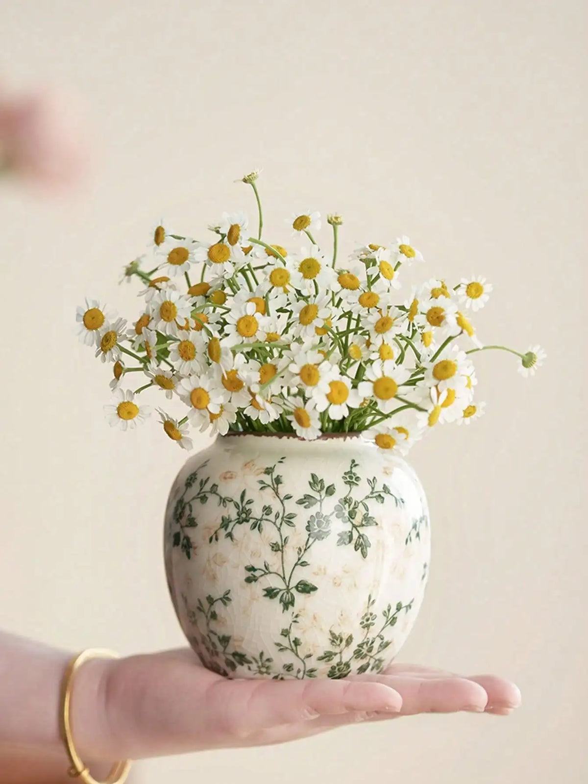 Eine Hand hält eine weiße Keramikvase mit gelben und weißen Wildblumen vor einem hellen Hintergrund.