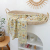 A woven basket holds a patterned muslin baby swaddle blanket draped over the edge of a white shelf.