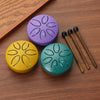 A set of three mini steel tongue drums in purple, yellow, and green with wooden mallets resting on a brown wooden surface.