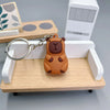 An adorable brown capybara keychain charm resting on a light wooden tray with a white rectangular box in the background.