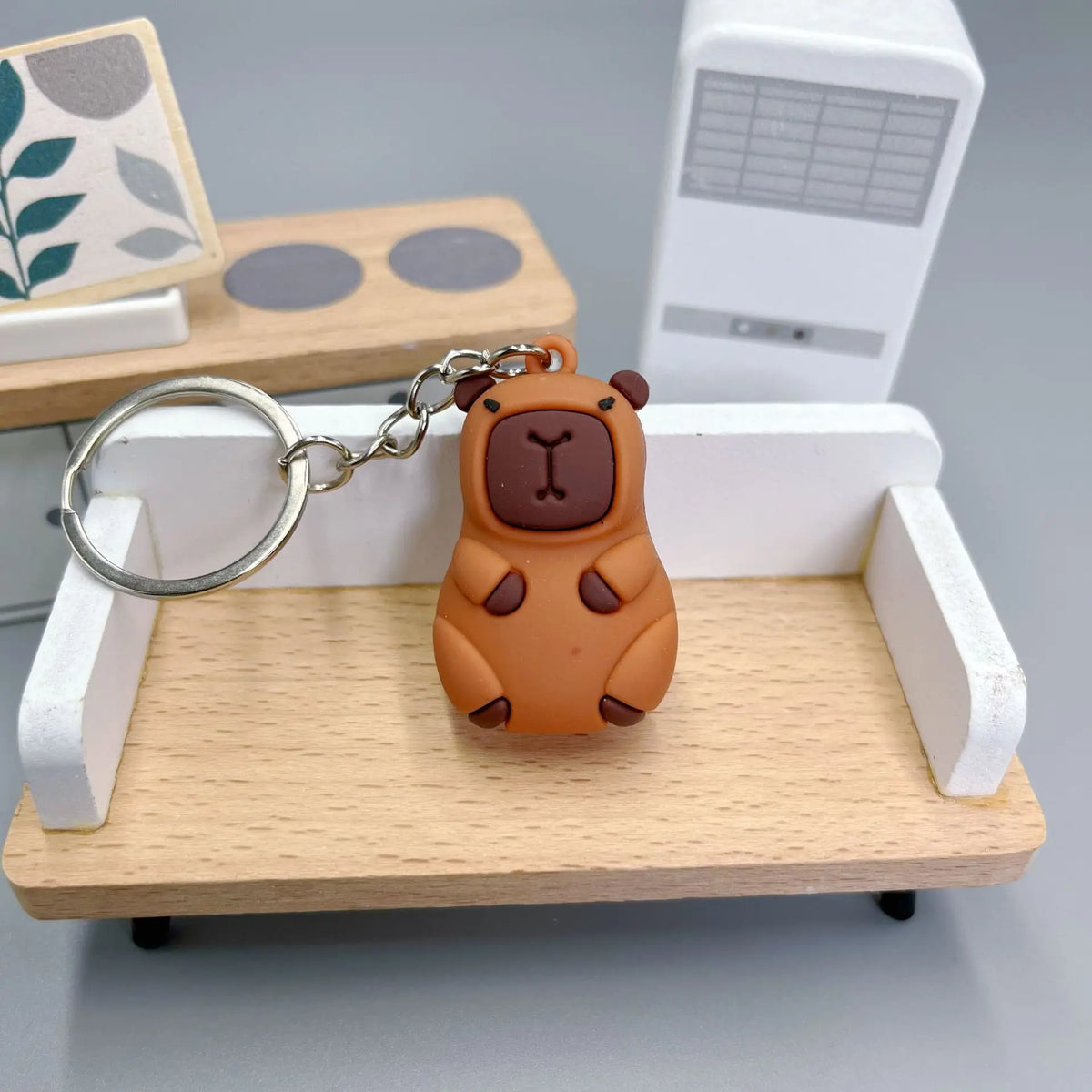 An adorable brown capybara keychain charm resting on a light wooden tray with a white rectangular box in the background.