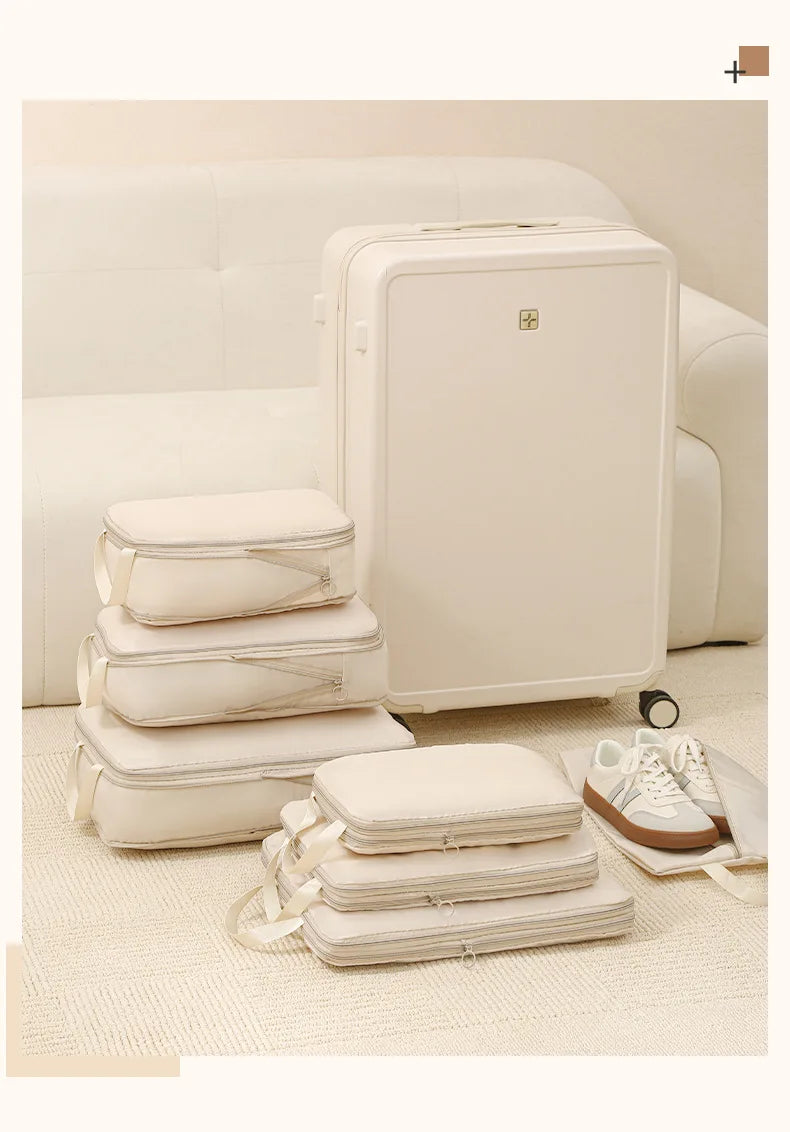 A set of white compressible packing cubes, including a large suitcase and several smaller bags, displayed on a light beige ca