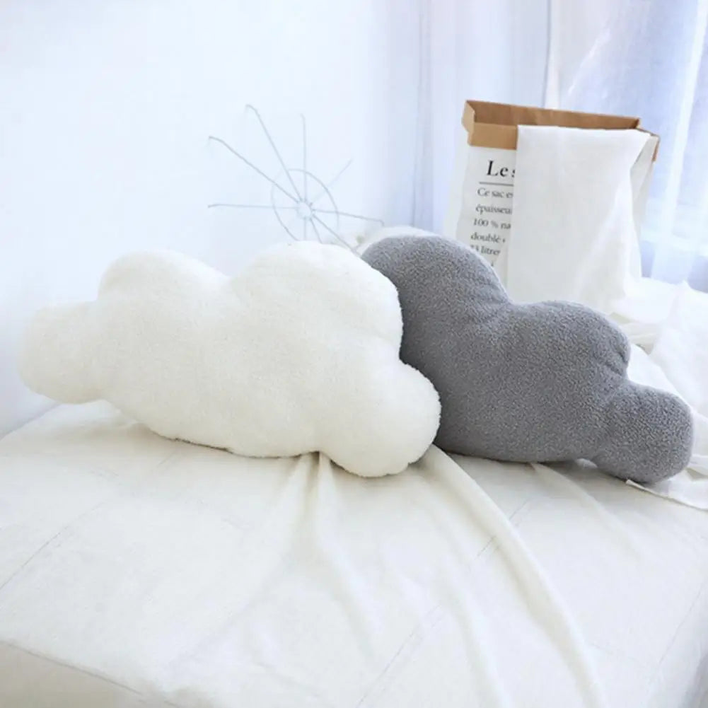 A white and gray cloud-shaped plush throw pillow resting on a white bed with a brown paper bag nearby.