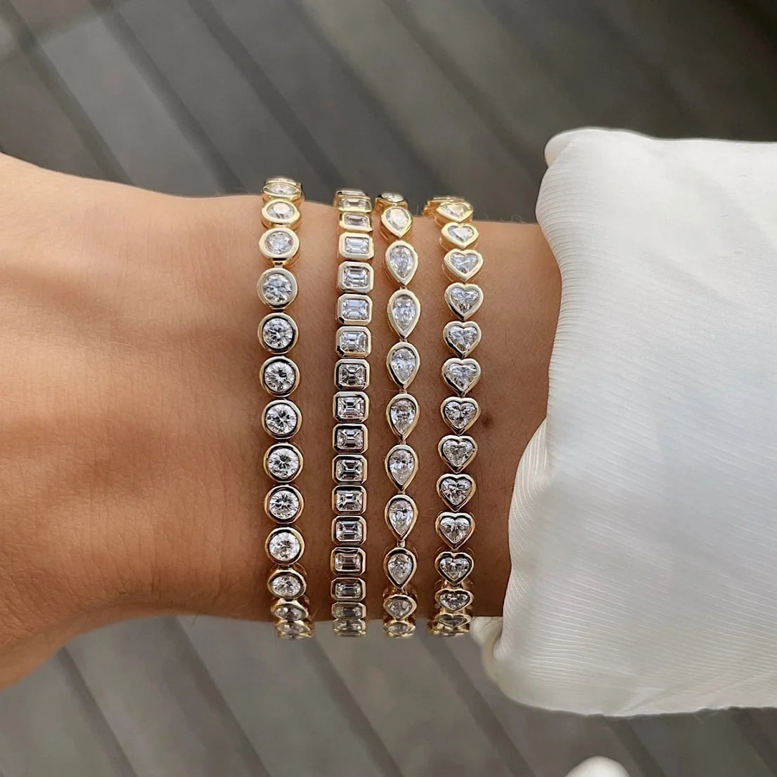 A close-up of a hand wearing a set of gold tennis bracelets featuring clear stones and heart-shaped accents.