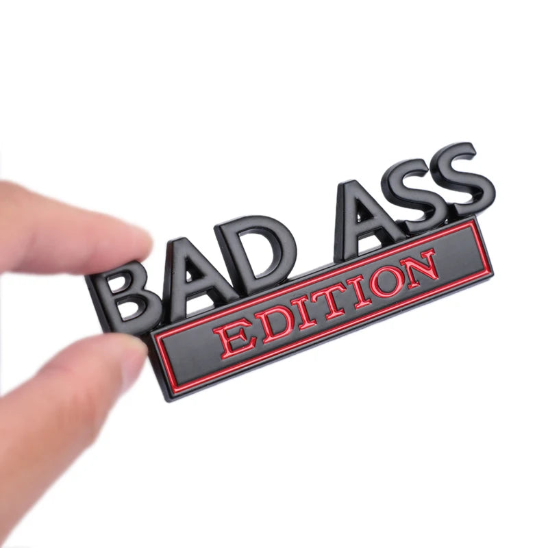 A black 3D metal badge emblem with the text BAD ASS EDITION is held by a hand.