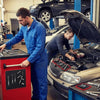 A mechanic in a blue jumpsuit works on a car engine with tools nearby.